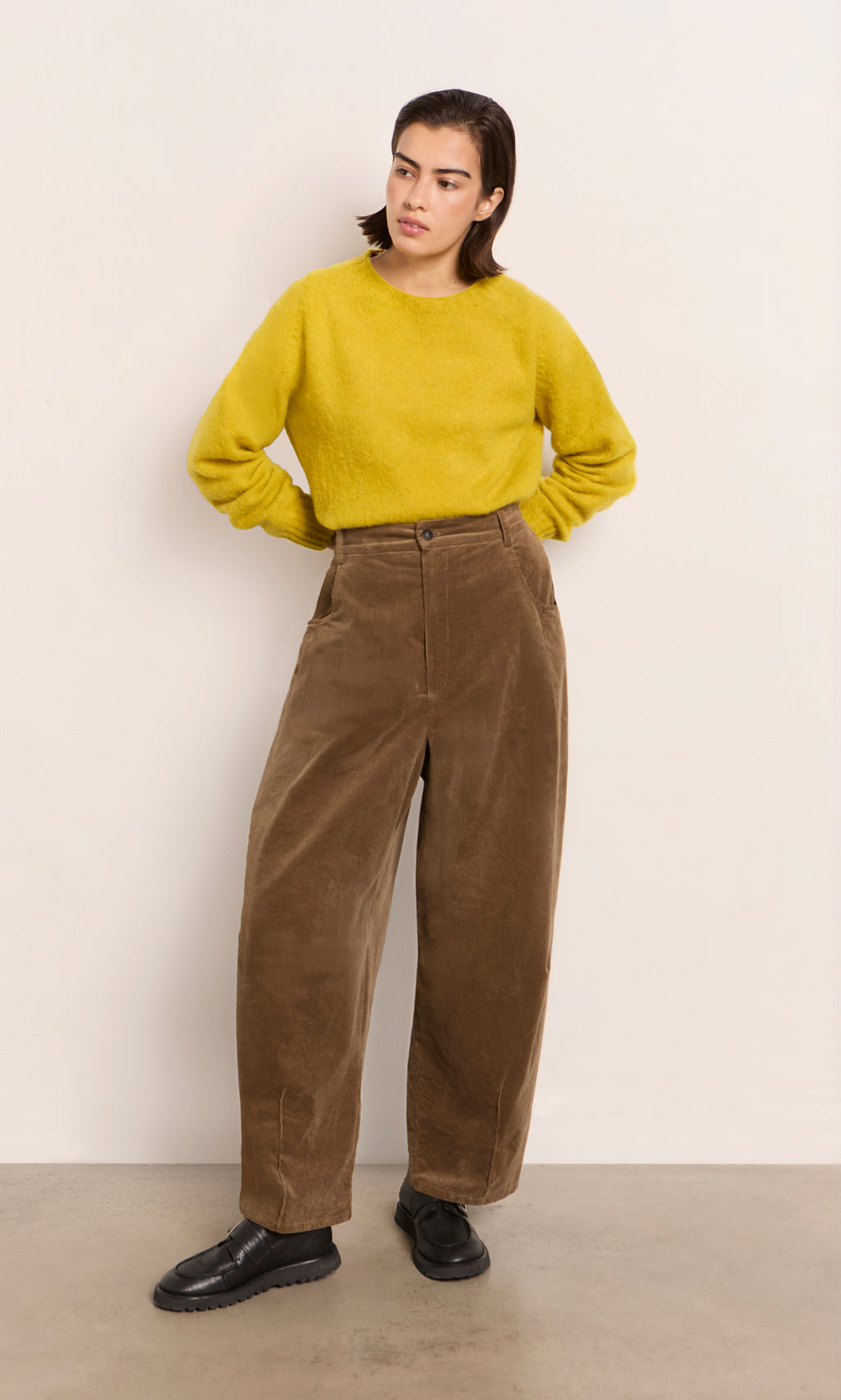 Plümo  Person in a yellow sweater, Breda Pants, and black shoes stands by a plain light wall.
