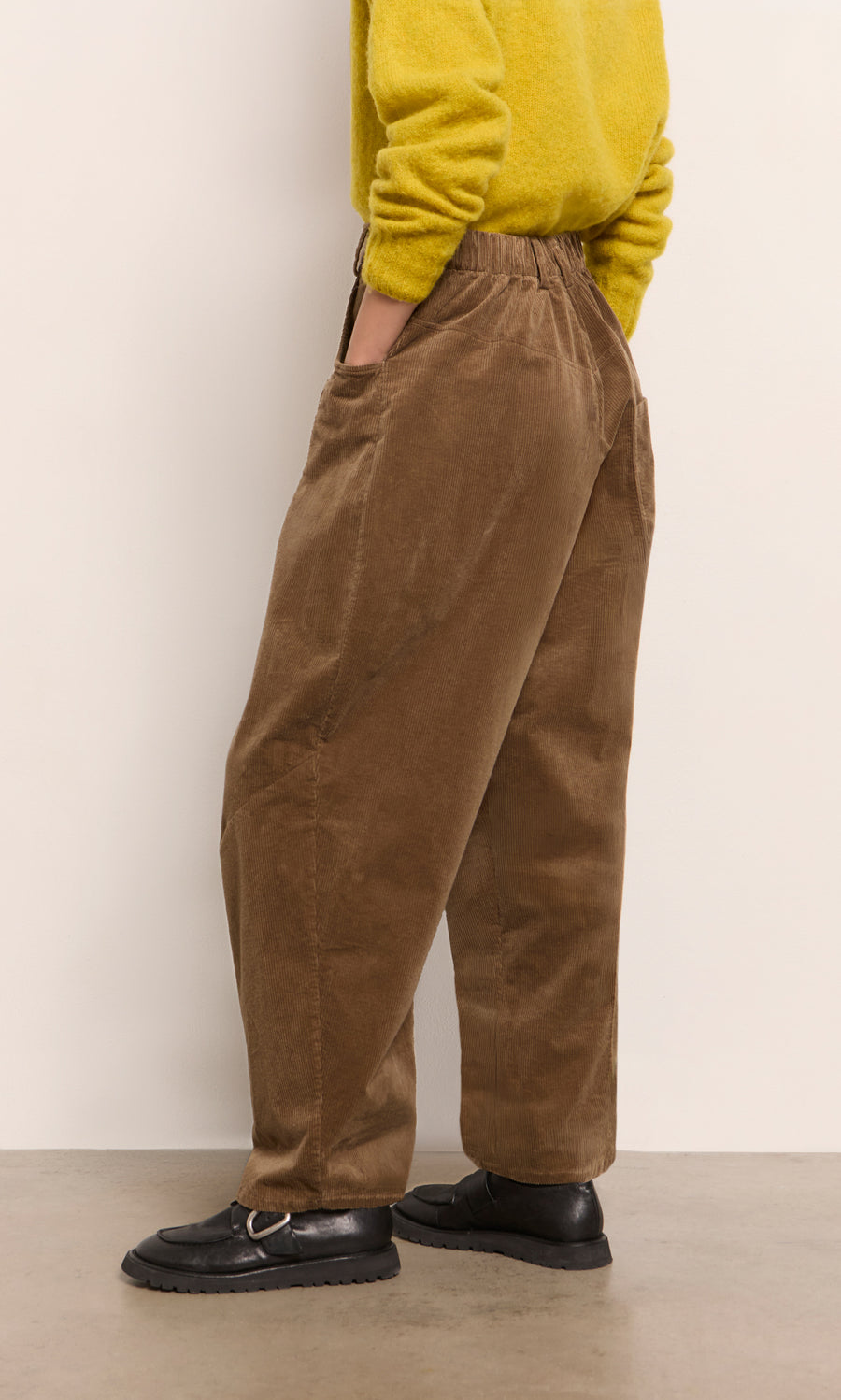 Plümo  Person in Breda Pants, yellow sweater, and black shoes stands sideways against a neutral background.