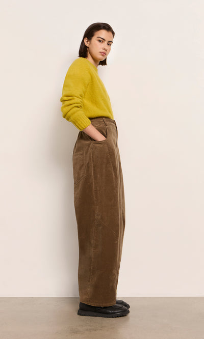 Plümo  Person in a yellow sweater and Breda Pants stands sideways with hands in pockets against a plain wall.