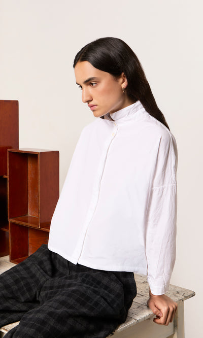 Plümo  Person in a Bressay Shirt and plaid pants sits on a table, looking to the side with a neutral expression.