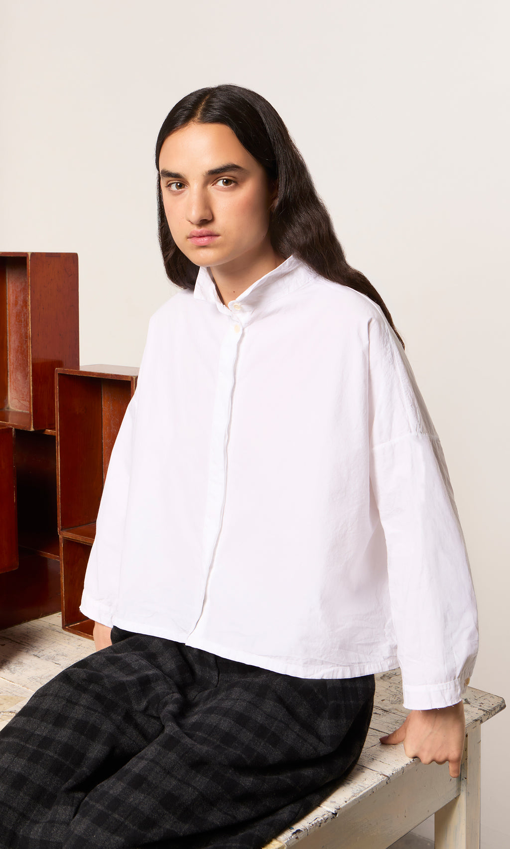 Plümo  Person wearing a white Bressay Shirt and plaid pants sits by wooden boxes on a light-colored table.