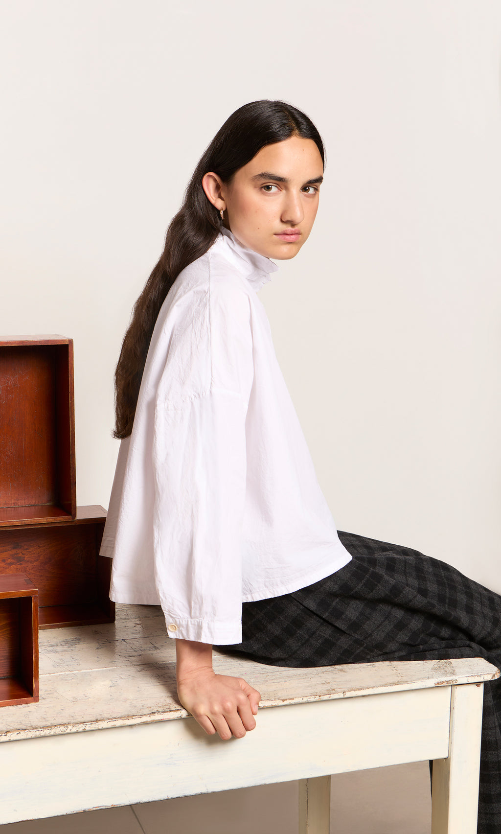 Plümo  Person with long dark hair in a relaxed Bressay Shirt sits sideways on a table, boxes beside them.