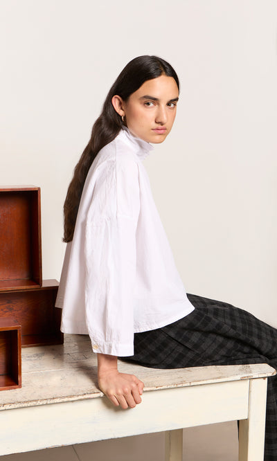 Plümo  Person with long dark hair in a relaxed Bressay Shirt sits sideways on a table, boxes beside them.