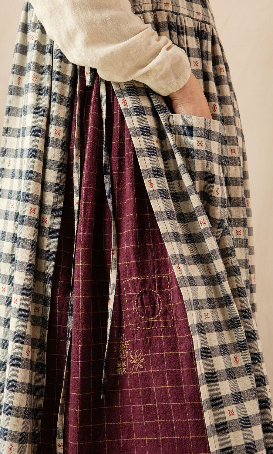 Plümo- A hand slips into the large pocket of the OMalley Apron Dress, a blue checked skirt with adjustable side ties.