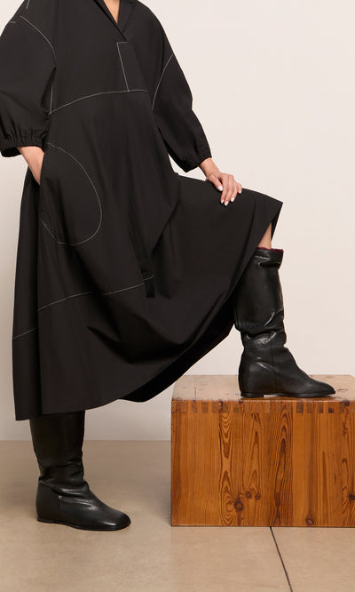 Plümo  Person in a loose black dress and Briony Boots rests one leg on a wooden block, hand on knee.