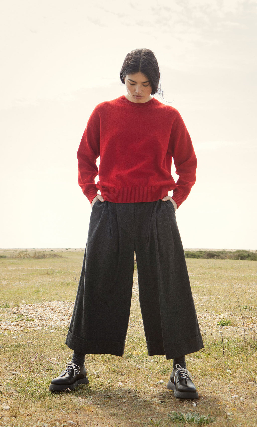 Plümo- Woman outdoors in a Bute Sweater, red crewneck jumper, and wide dark pants, looking down with hands in pockets.