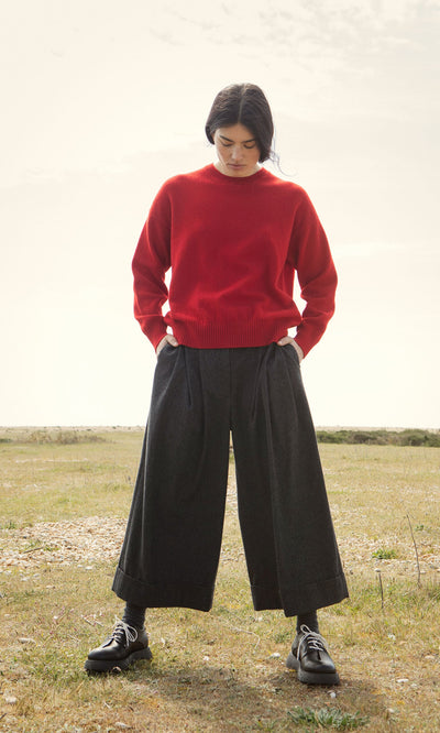 Plümo- Woman outdoors in a Bute Sweater, red crewneck jumper, and wide dark pants, looking down with hands in pockets.