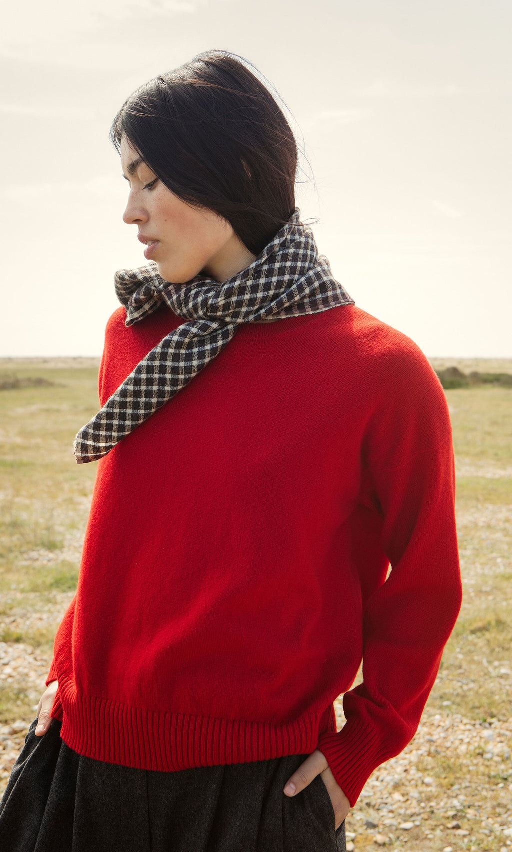 Plümo- Woman wears a Bute Sweater and plaid scarf outdoors, hands in pockets, with a grassy field in the background.
