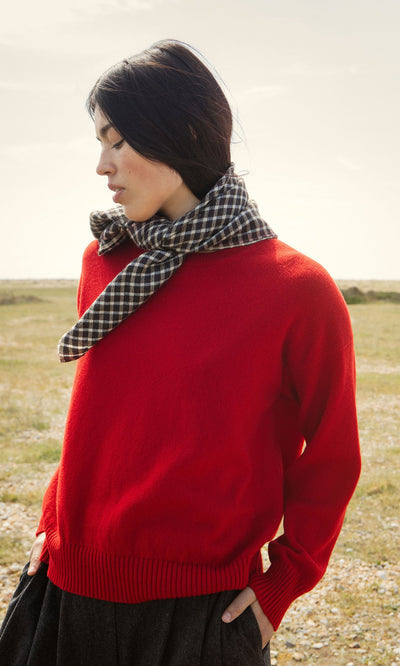 Plümo- Woman wears a Bute Sweater and plaid scarf outdoors, hands in pockets, with a grassy field in the background.