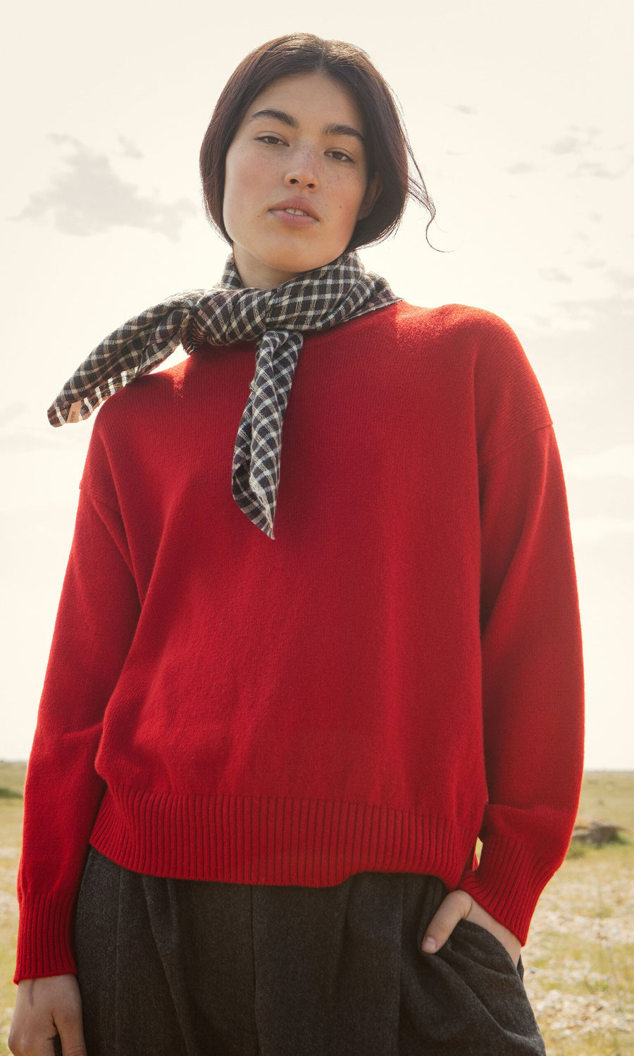 Plümo- Person wearing a Bute Sweater, plaid scarf, and dark pants stands outdoors with a neutral expression.