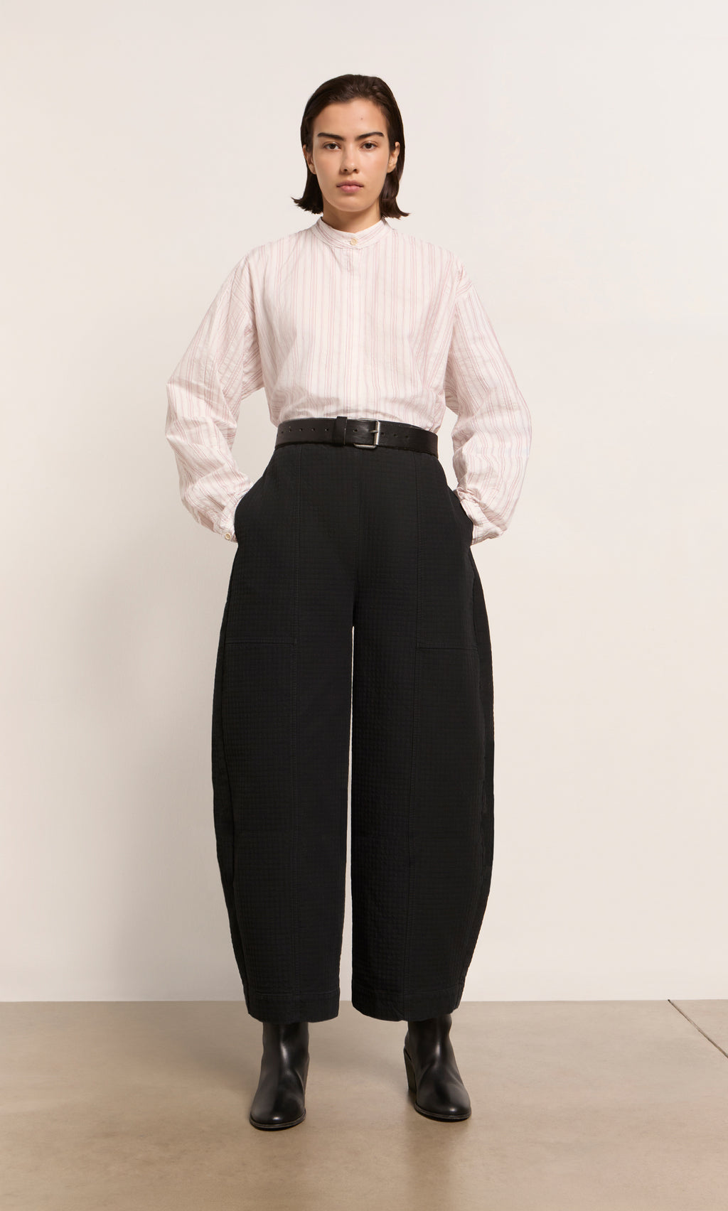 Plümo  A person wears a light striped shirt, Calow Pants - Coal with a wide black belt, and black boots against a plain background.