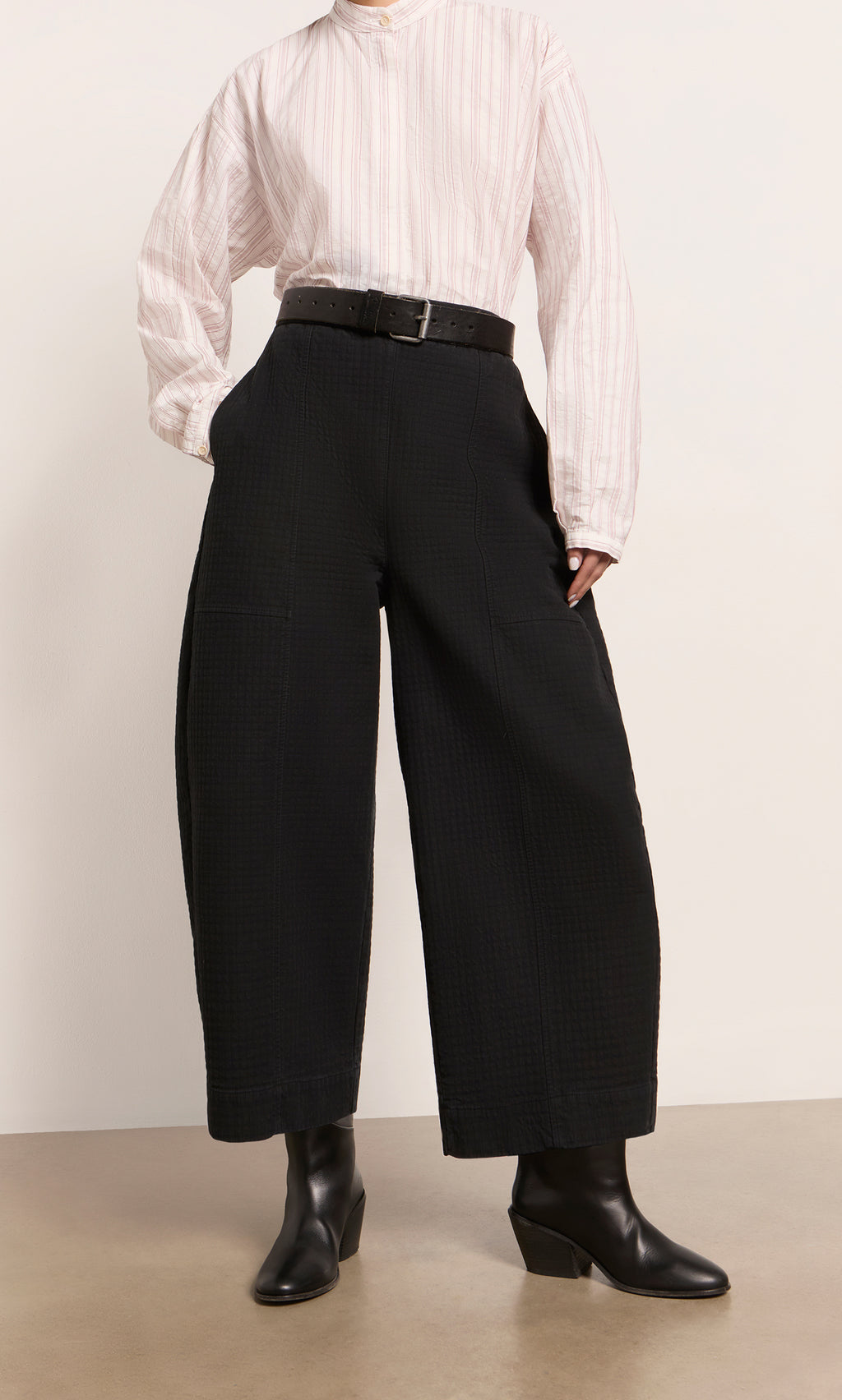Plümo  Person in a pink striped shirt, Calow Pants - Coal, black belt, and black heeled boots standing indoors.