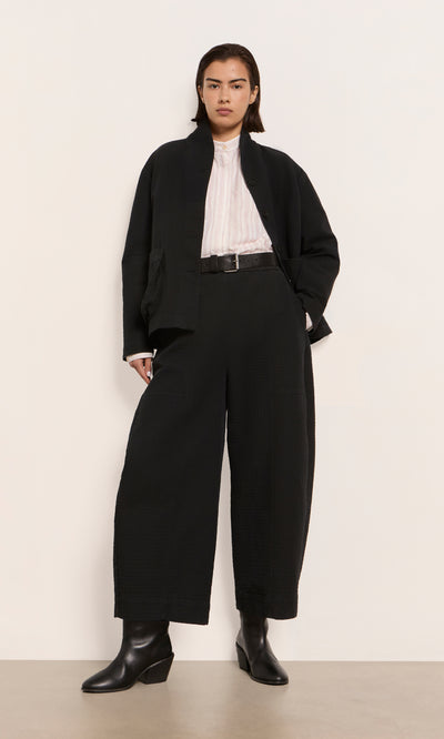 Plümo  Person in Calow Pants - Coal with a black jacket, white top, black belt, and boots, standing by a plain wall.