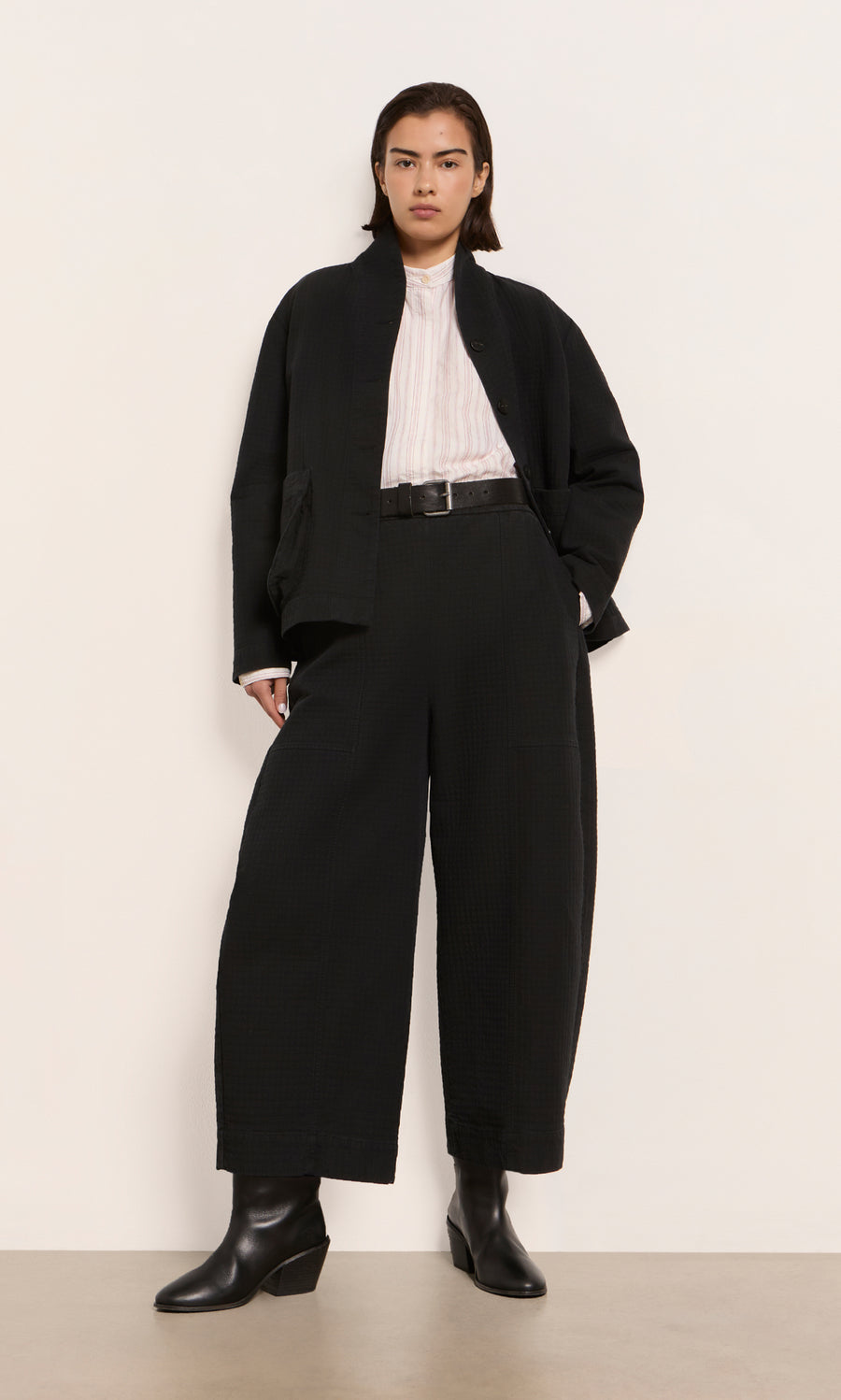 Plümo  Person in Calow Pants - Coal with a black jacket, white top, black belt, and boots, standing by a plain wall.