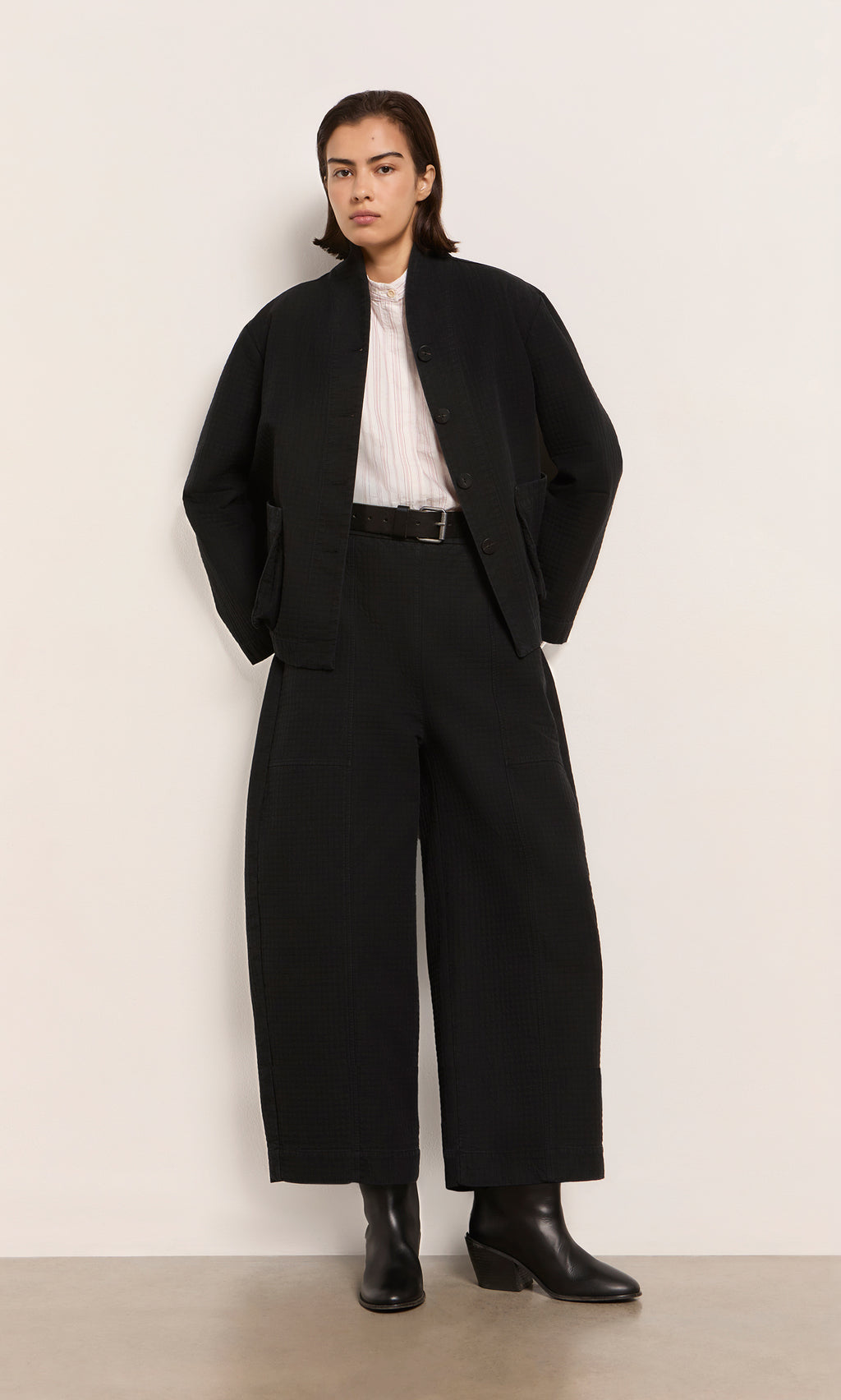 Plümo  Person wears Calow Pants - Coal, black jacket, black boots, and white shirt, standing against a plain background.
