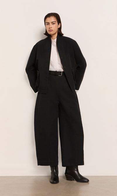 Plümo  Person in Envoy Jacket - Coal, wide-leg pants, white shirt, black boots—utilitarian style designed in London.