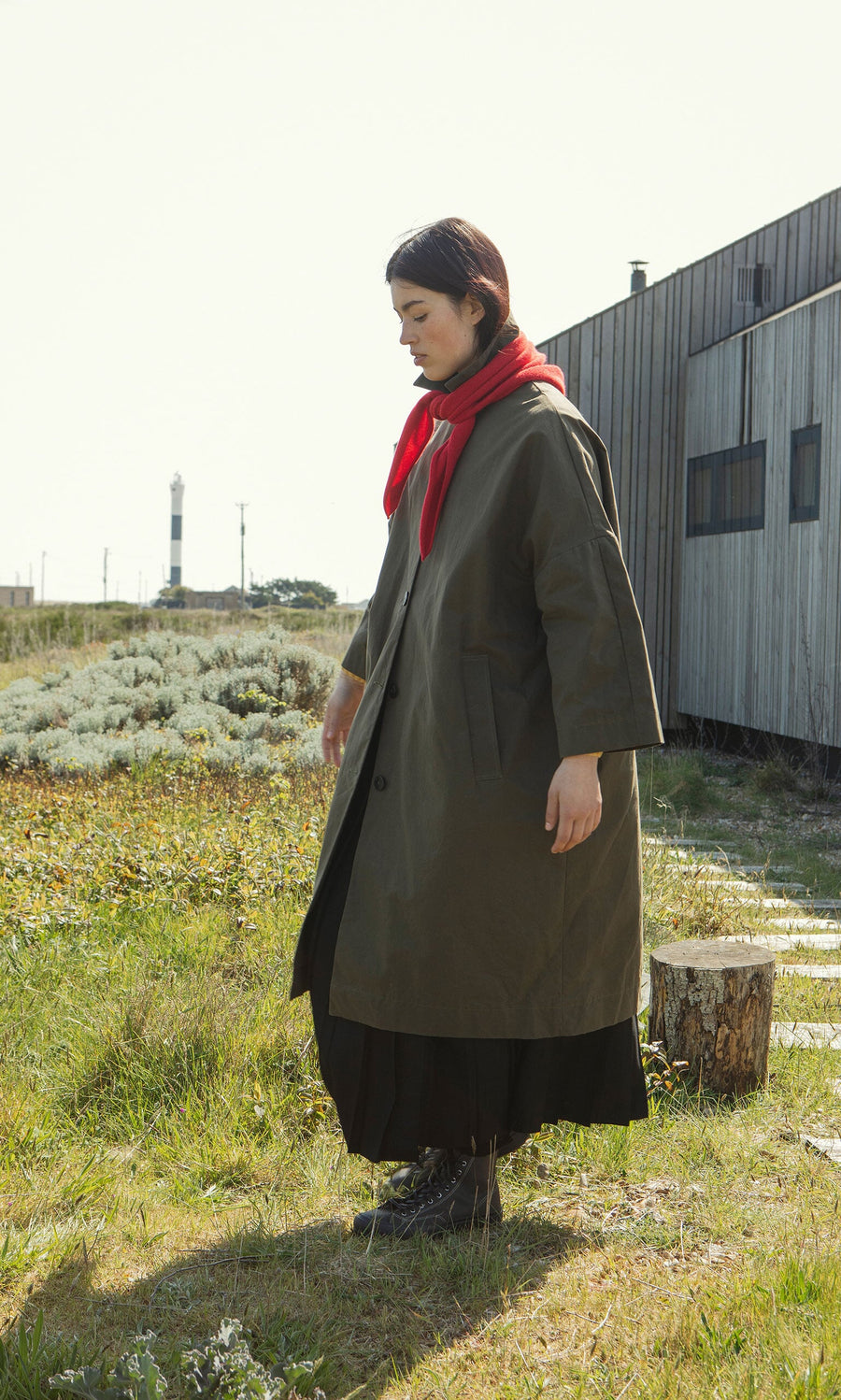 Plümo- Person in a Doherty Coat - Olive and red scarf stands on grass near a modern wooden building and lighthouse.
