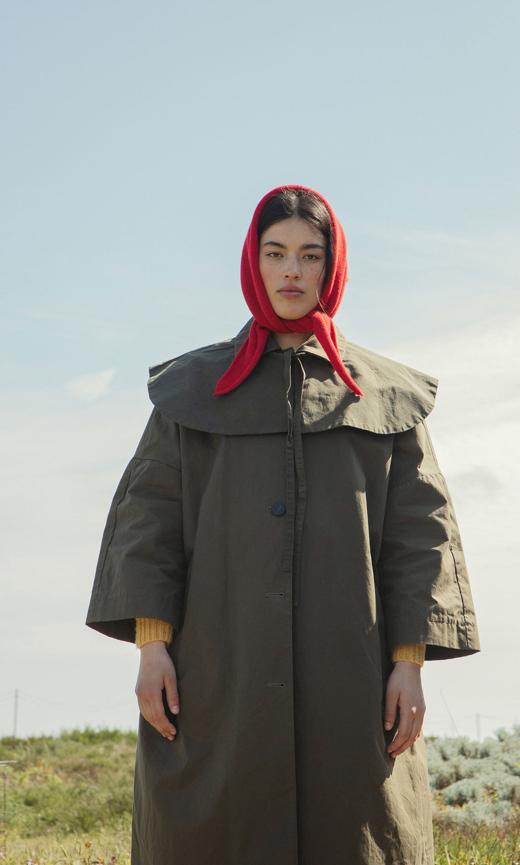 Plümo- Person wearing the Cape Collar olive green waxed cotton coat and red headscarf stands outdoors, looking at the camera.