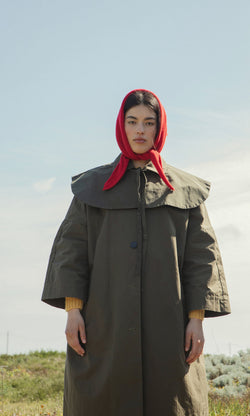 Plümo- Person wearing the Cape Collar olive green waxed cotton coat and red headscarf stands outdoors, looking at the camera.