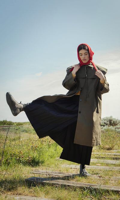 Plümo- A woman in a red headscarf and Doherty Coat - Olive lifts one foot on a grassy path outdoors.