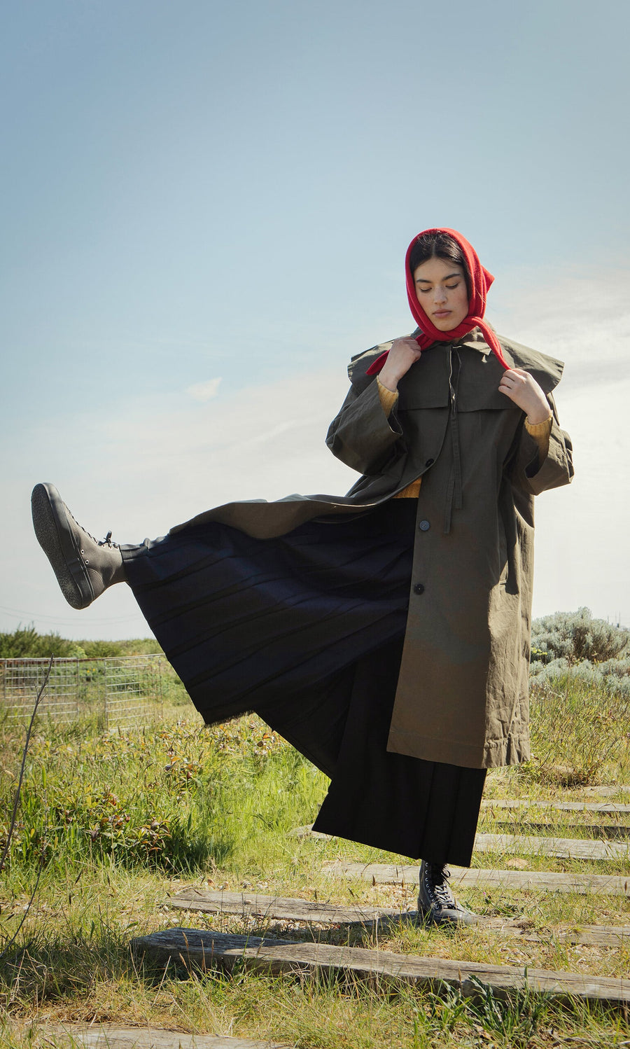 Plümo- A person in a red headscarf and long coat with a Cape Collar lifts one leg on a grassy path under a blue sky.