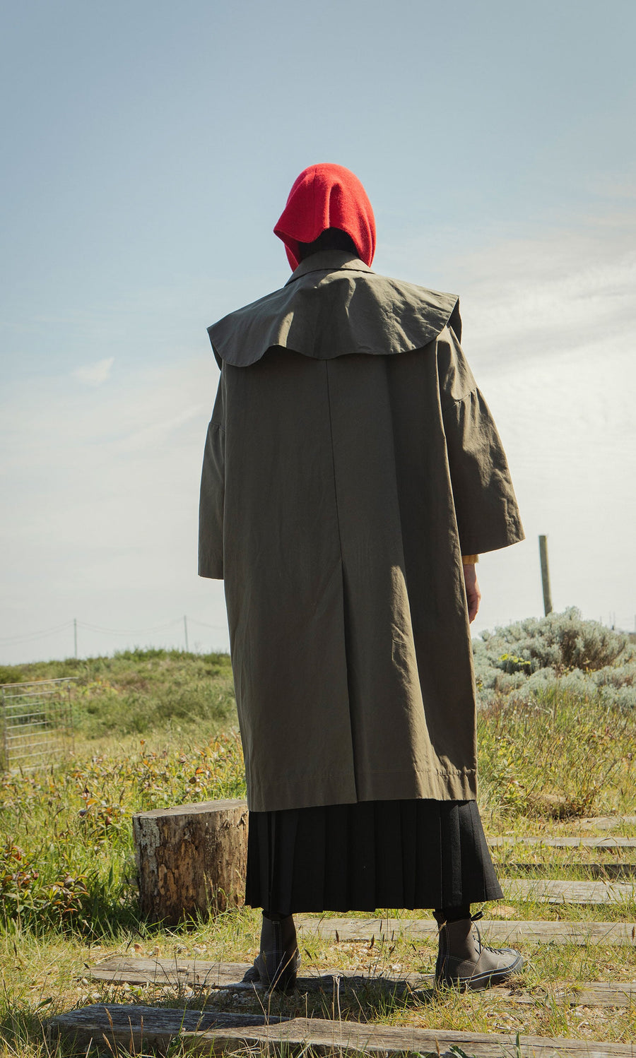 Plümo- A person in a long olive Cape Collar coat with a red hood stands outdoors on a grassy path, facing away.