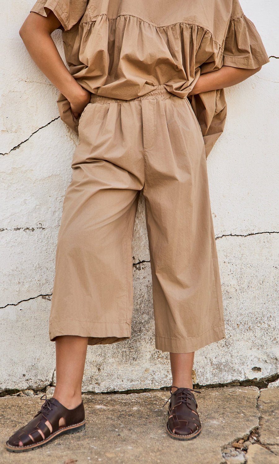 Plümo- Person in loose tan top and Howland Pants with brown sandals, standing against a cracked white wall.