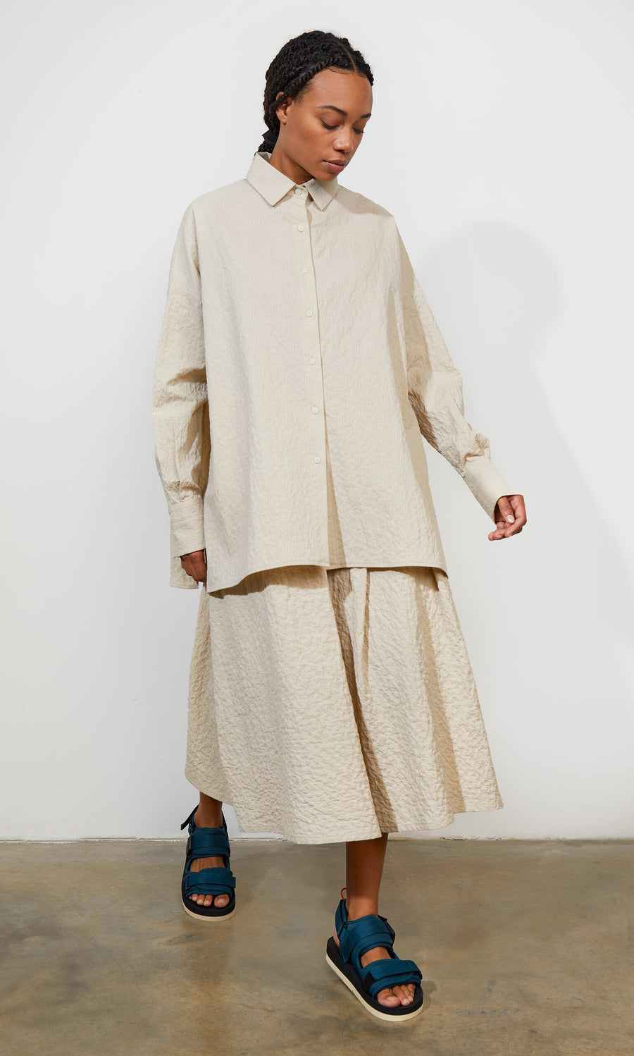 Plümo- A woman in a beige shirt and Jane Skirt with teal sandals walks indoors on a concrete floor.