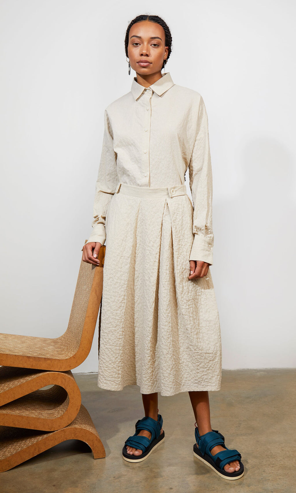 Plümo- Woman in a beige textured shirt and Jane Skirt set with teal sandals, by a sculptural wooden chair.