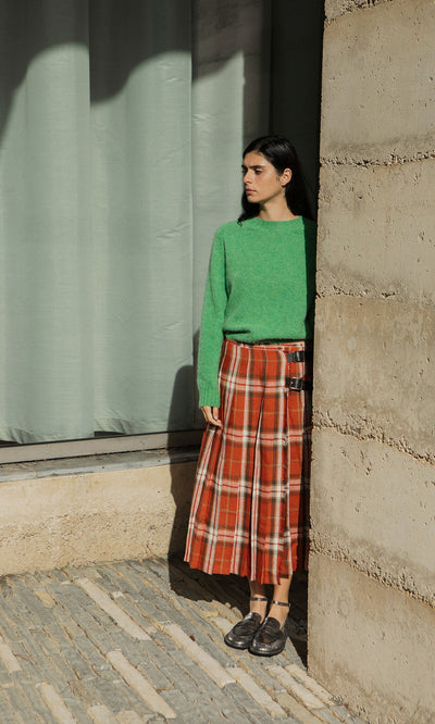 Plümo- Woman in green sweater and Clemie Kilt with leather buckle stands by a concrete wall, partly in shadow, looking right.