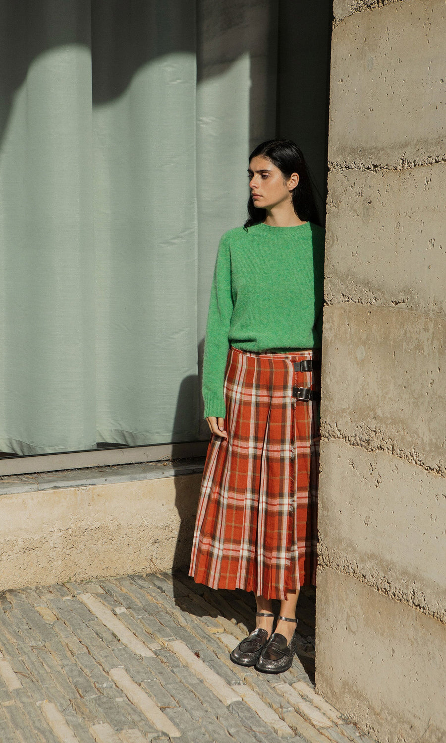 Plümo- Woman in green sweater and Clemie Kilt with leather buckle stands by a concrete wall, partly in shadow, looking right.