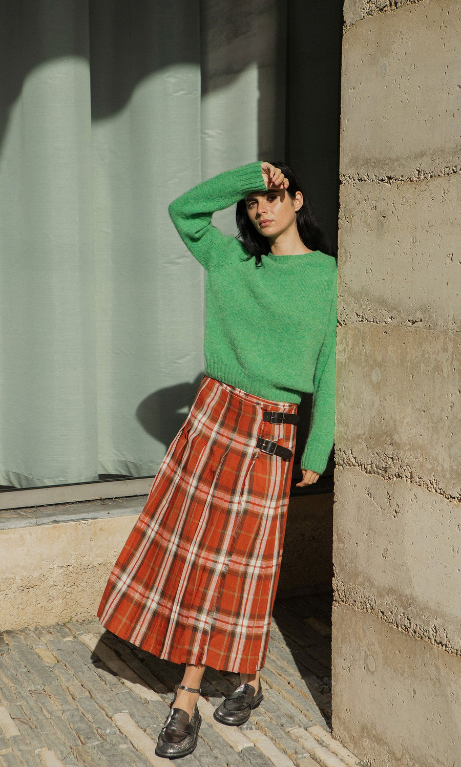 Plümo- Woman in green sweater and Clemie Kilt leans against a wall, shielding her eyes from the sun.
