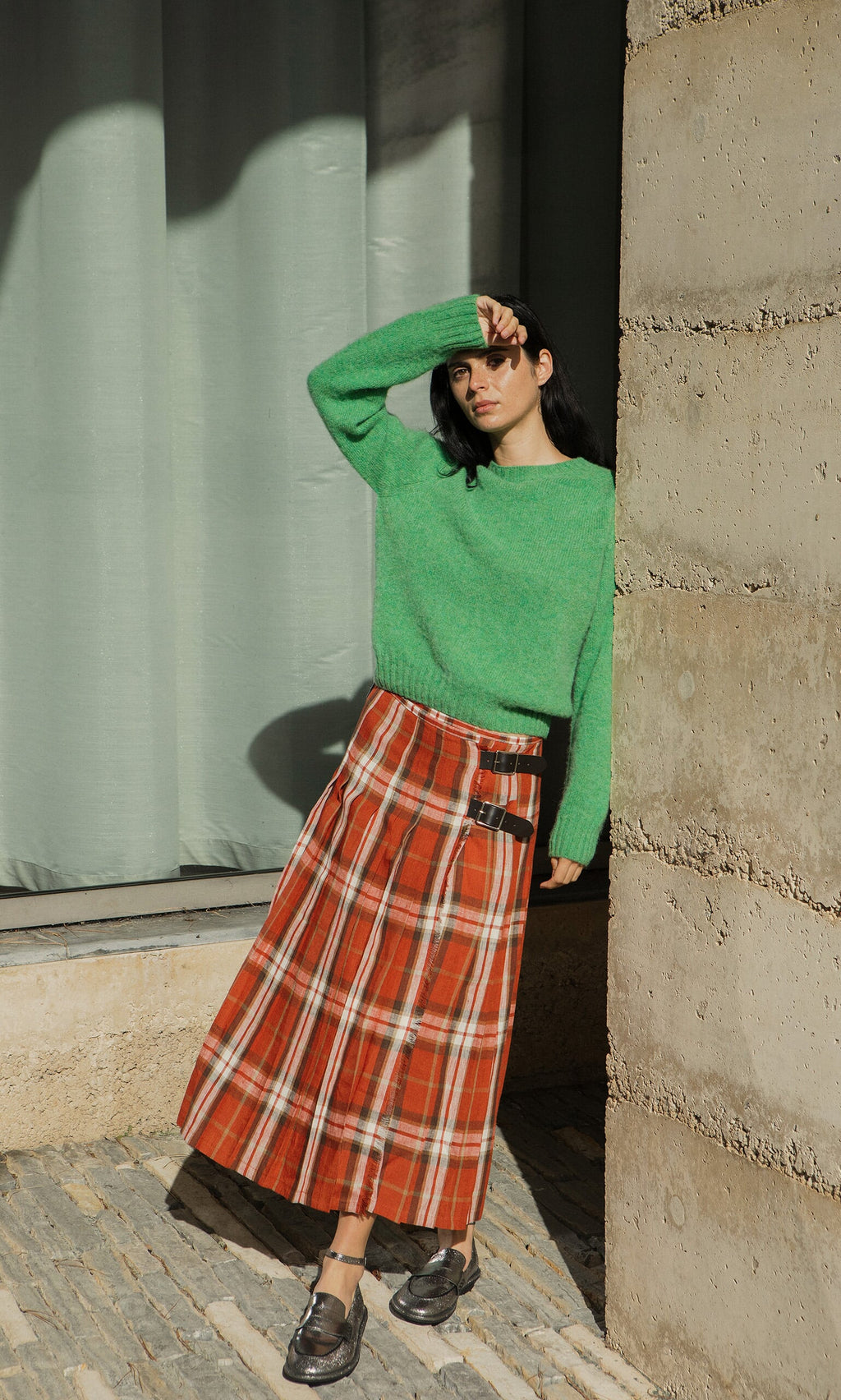 Plümo- Woman wearing the Wicklow Crewneck - Garden Leaf leans on a stone wall, shielding her eyes from the sun.