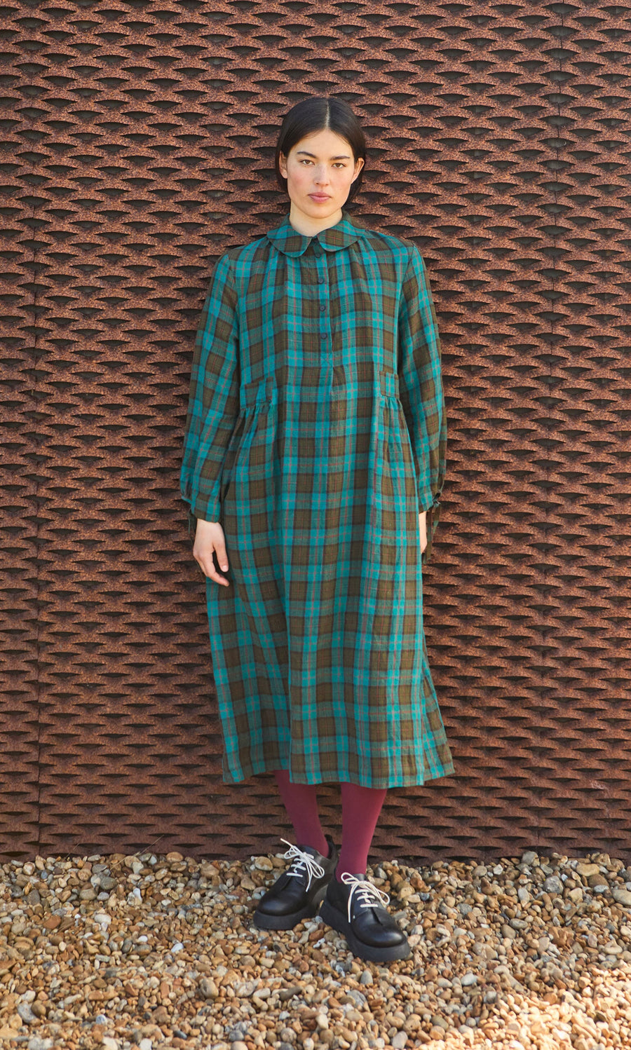 Plümo- Woman stands on pebbles in front of a textured wall, wearing the Clodagh Tartan Dress with maroon tights.