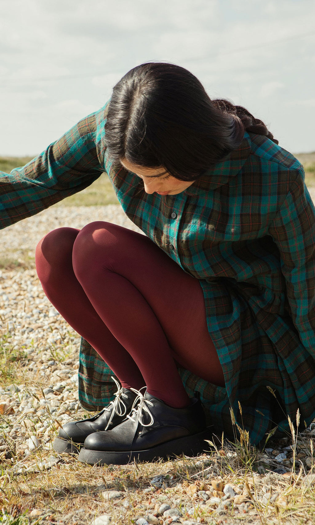 Plümo- A person in a green plaid dress and Deep Red Tights from Sweden squats outdoors on rocky ground, looking down.