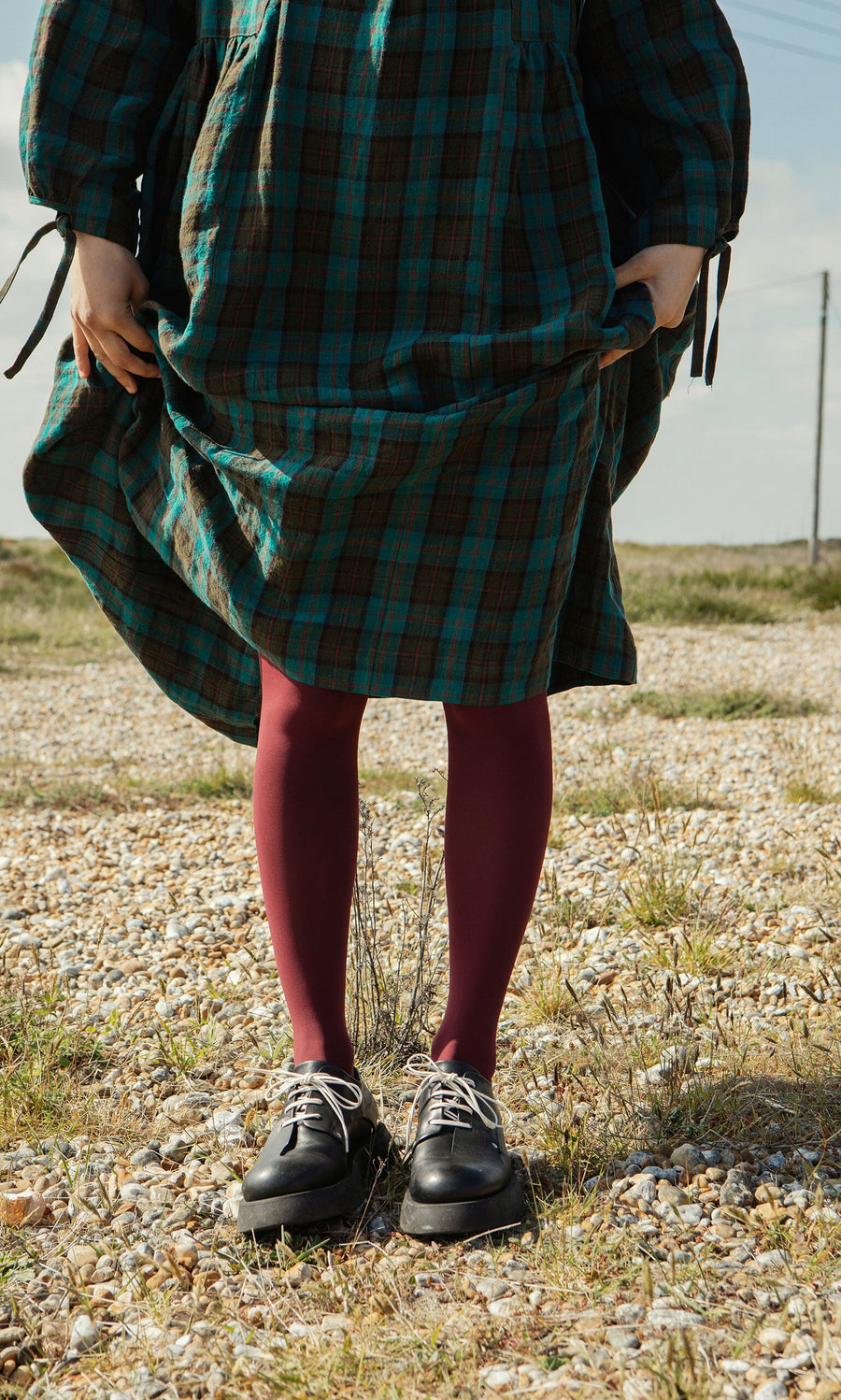 Plümo- Person in a green plaid dress, maroon tights, and Hosier Brogues stands on a gravel path, holding their dress.