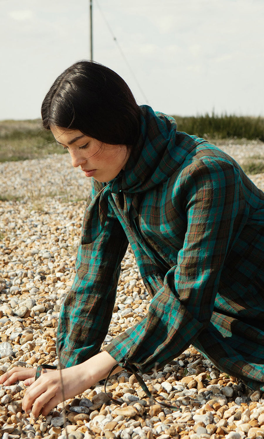 Plümo- Woman in the Clodagh Tartan Dress with a Peter Pan collar kneels on rocks, gently touching the stones.