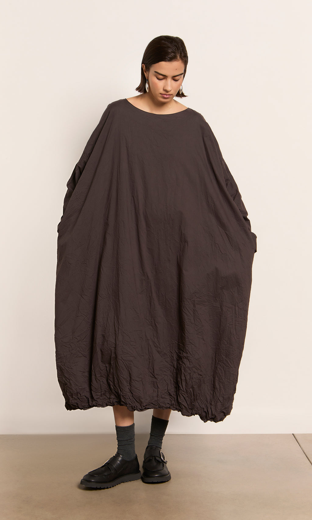 Plümo  Person in a loose, peaty brown Cloud Dress, black shoes, gray socks, looking down.