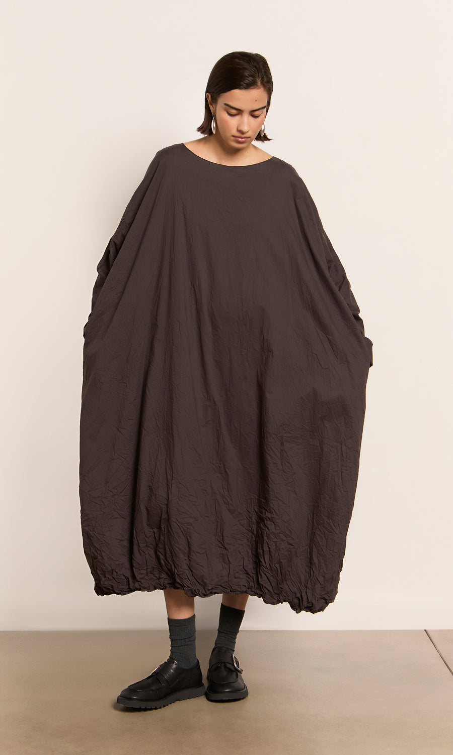 Plümo  Person in a loose, peaty brown Cloud Dress, black shoes, gray socks, looking down.