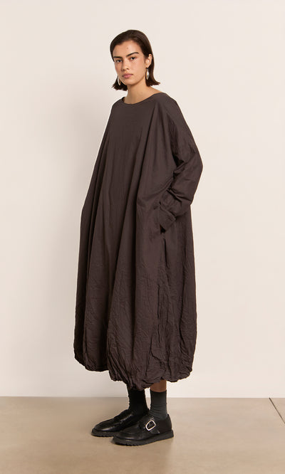 Plümo  Person in the Cloud Dress, a peaty brown bubble-hem maxi, with black shoes stands before a light plain backdrop.