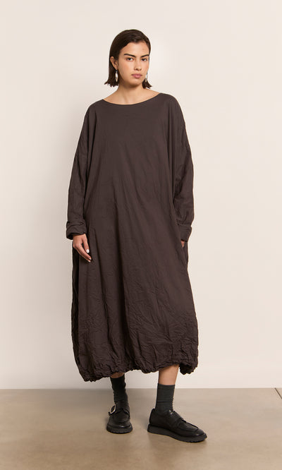 Plümo  Person wears the Cloud Dress, a loose peaty brown maxi with bubble hem, plus black shoes and socks, on a plain backdrop.