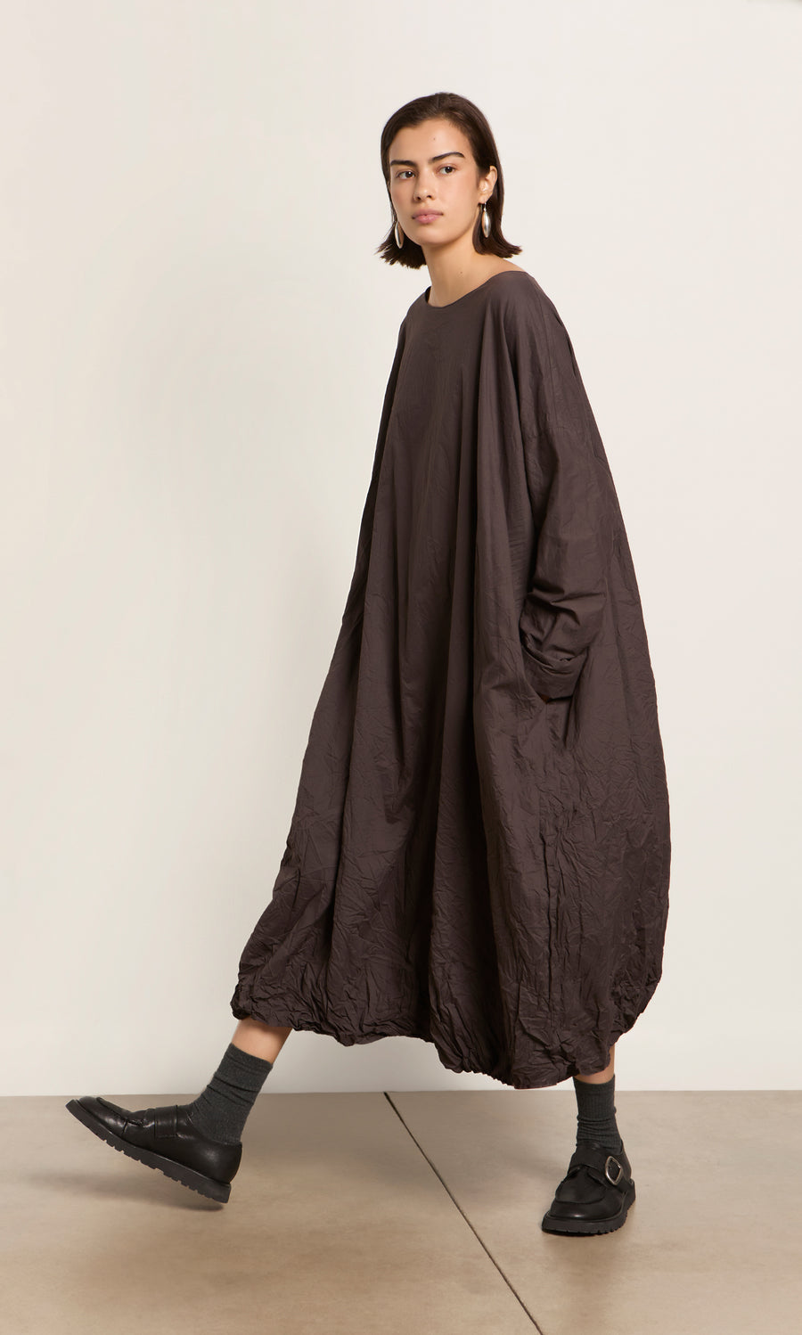 Plümo  Person in a loose, long, dark brown dress with pockets, gray socks, and Black Creepers walking indoors.
