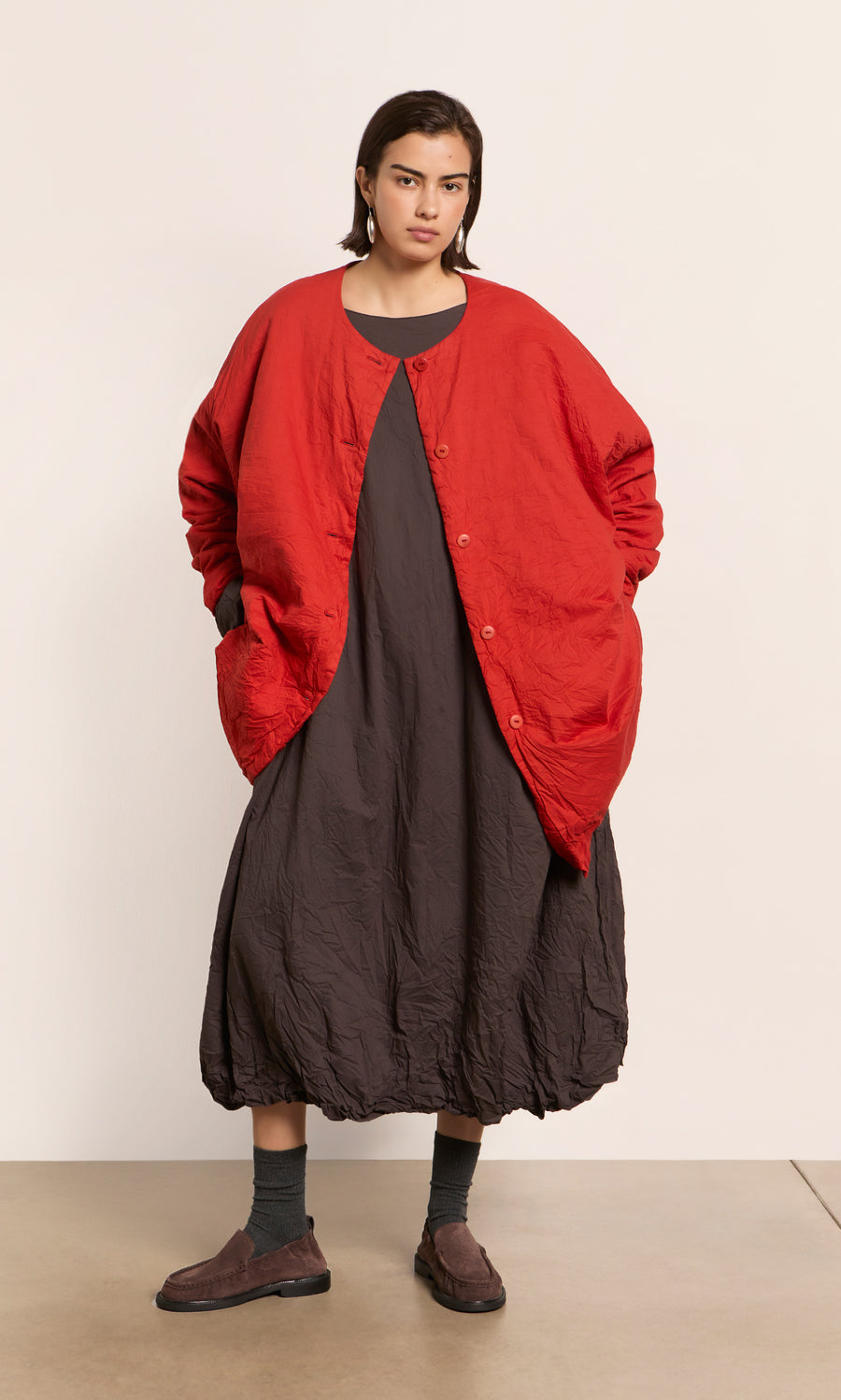 Plümo  Person wears the Cloud Dress—a peaty brown bubble-hem maxi—with a loose red jacket, hands in pockets.