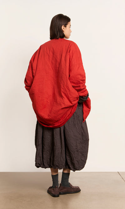 Plümo  Person standing sideways in a hand-dyed cotton red Coburn Jacket, baggy skirt, socks, and brown shoes on plain background.