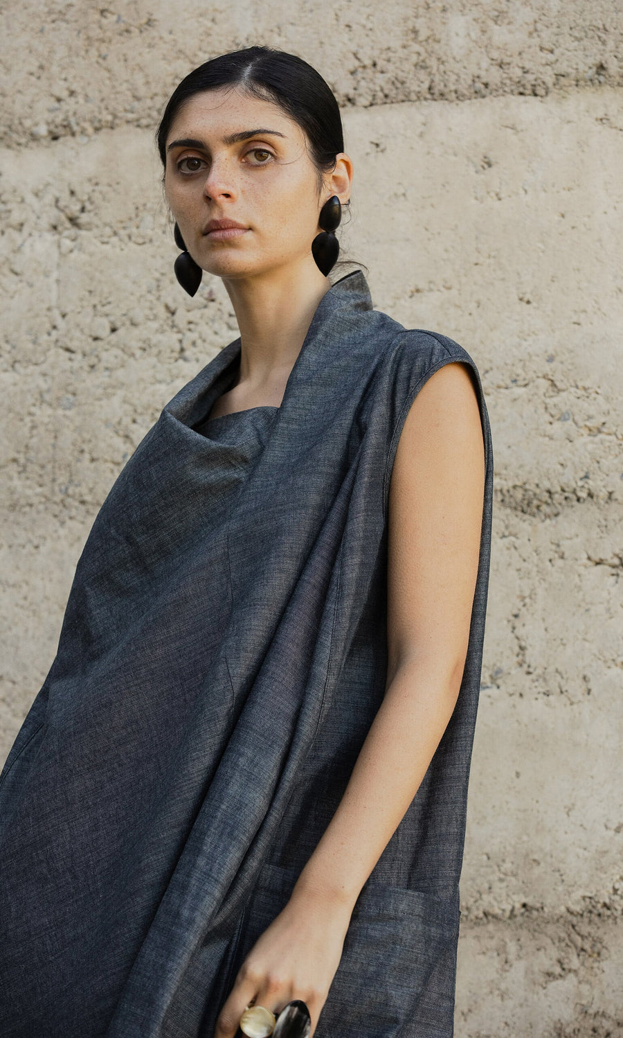 Plümo- Woman in the Emi Dress, a sleeveless gray asymmetric dress, wears black earrings and stands before a concrete wall.