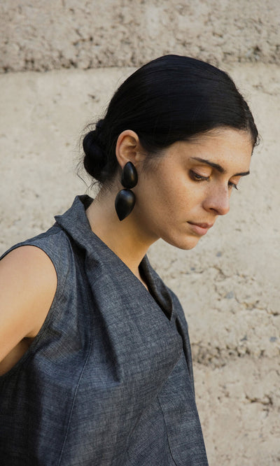 Plümo- Woman with dark hair in a low bun, wearing Coalvale Earrings and a gray sleeveless top, stands by a textured wall.