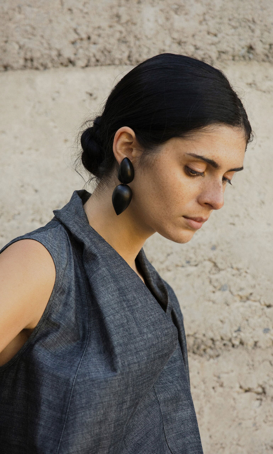 Plümo- Woman with dark hair in a low bun, wearing Coalvale Earrings and a gray sleeveless top, stands by a textured wall.