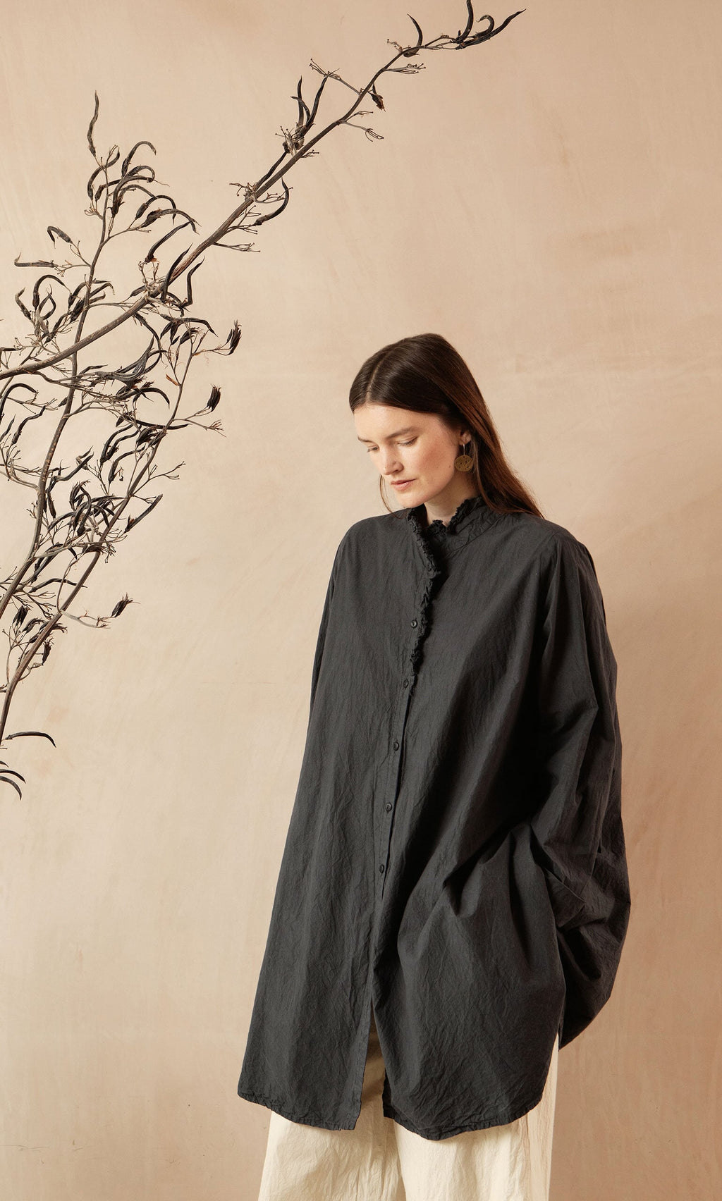 Plümo- Woman in the Cockle Shirt stands beside a thin, leafless branch against a beige background.