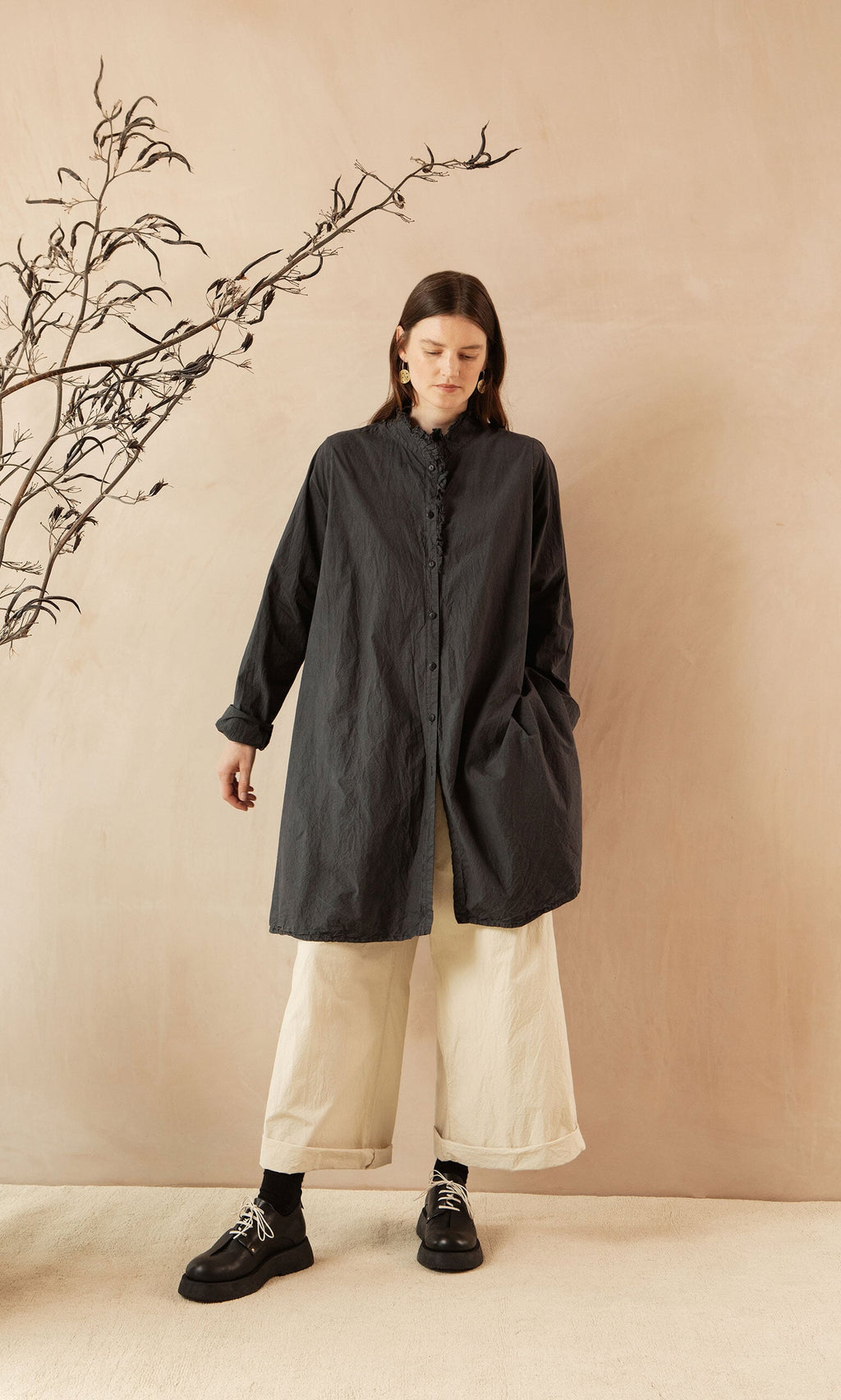 Plümo- Woman in a black Cockle Shirt, wide cream pants, and boots stands against a beige background with a branch.
