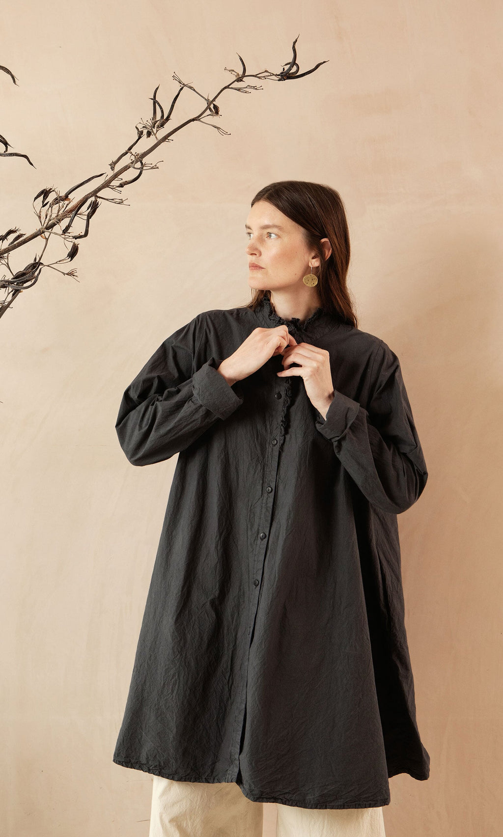 Plümo- Woman in a long, loose Cockle Shirt stands against a beige wall with a branch in the background.